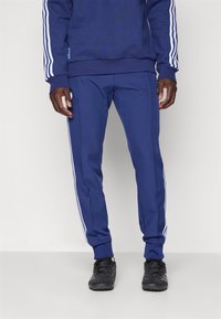 Tumši zili sporta bikses ar baltām sānu svītrām, elastīgu jostu, ribveida aprocēm un mīkstu audumu. Kombinētas ar melniem sporta apaviem.