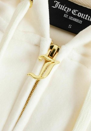 Witte Juicy Couture hoodie met gouden ritssluiting in de vorm van een gestileerde letter "J" en zichtbaar merkembleem aan de binnenkant van de kraag.