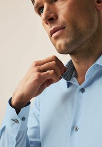 Chemise bleu clair avec un tissu texturé, un léger éclat et un col à motifs ; présente des poignets à boutons et un gros plan d'une main ajustant le col.