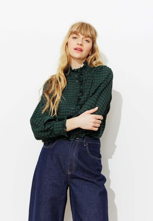 Femme blonde aux longs cheveux portant un chemisier à carreaux vert foncé avec des volants et un jean bleu large taille haute, posant les bras croisés.