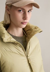 Ανοιχτό ελαιόπετσο πνευματισμένο μπουφάν με ψηλό γιακά, κλείσιμο με φερμουάρ και μαλακή υφή. Φορεμένο πάνω από ένα γκρι πουλόβερ, σε συνδυασμό με ένα μπεζ καπέλο.