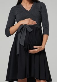 Těhotenské šaty s šedým vrškem a černou sukní. Zavinovací design s třičtvrtinovými rukávy a sepnutým pasem, vyrobené z měkké tkaniny.