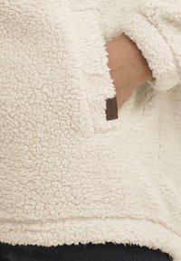 Kreemit värvi fliispullover, millel on tekstureeritud, karvane pind. Omab taskut ja väikest pruuni nahasildi külgedel.