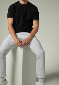 Camiseta negra de manga corta con textura y ribete gris en contraste, combinada con pantalones blancos y zapatillas blancas, sentada sobre un pedestal claro.