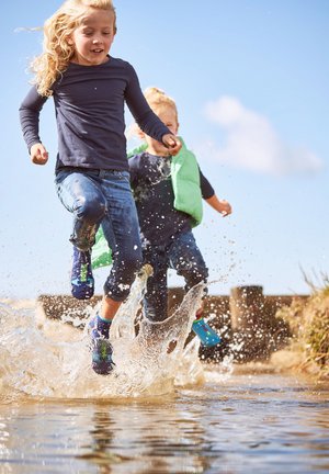 Two children jumping and splashing in a water puddle outdoors on a sunny day, wearing casual clothes and water shoes.