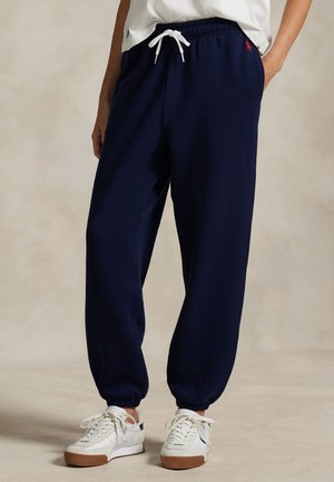 Person iført mørkeblå joggingbukser med elastiske ankler, hvid snøre og hvide sneakers med gummisåler, stående på en neutral baggrund.