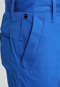 Modré kalhoty z hladkého materiálu, se skládáním, horními a bočními kapsami a logem na přední straně.