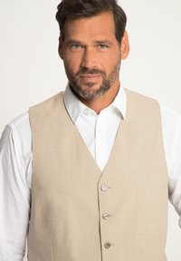 Beige Weste aus strukturiertem Stoff mit V-Ausschnitt, drei Knöpfen auf der Vorderseite, getragen über einem weißen Hemd mit Knöpfen.