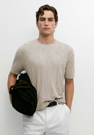 Giovane uomo con capelli mossi che indossa un maglione beige corto a maniche corte con trama e pantaloni bianchi tiene una grande borsa nera davanti a uno sfondo bianco uniforme.