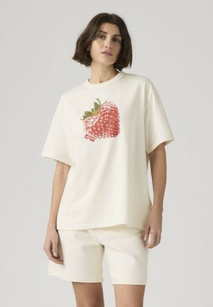 Kvinde iført off-white oversized t-shirt med stort rødt jordbærmotiv og matchende off-white shorts mod en ensfarvet baggrund.