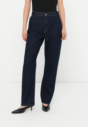 Jeans bleu foncé à taille haute et à jambes larges, avec une fermeture à bouton sur le devant et des poches. Portés avec des escarpins noirs à bouts pointus.