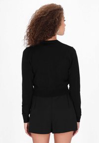 Schwarzer Strickpullover mit langen Ärmeln, gerippter Taille und Rundhalsausschnitt. Das Modell trägt schwarze Shorts.