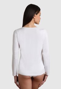 Donna con lunghi capelli scuri, che indossa un top bianco a maniche lunghe aderente e biancheria intima bianca, voltata di spalle contro uno sfondo chiaro e semplice.