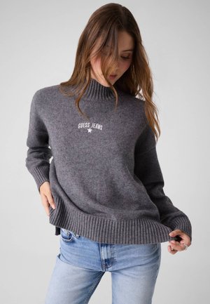 Frau trägt einen grauen Guess Jeans Pullover mit gerippten Bündchen und Kragen, kombiniert mit hellblauen Jeans, vor einem einfarbigen hellen Hintergrund.