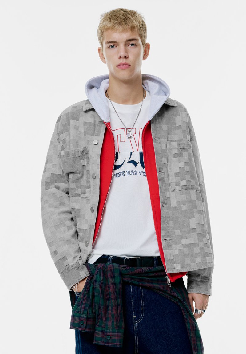 Grå mønstret jakke over en rød hættetrøje, båret med en hvid grafisk t-shirt. Blå jeans og en grønternet skjorte knyttet om taljen. Sølvtilbehør.