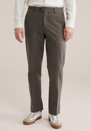 Mees, kes kannab pruune kohandatud pükse, valgeid tossusid ja valget pikkade varrukatega särki, millel on õhukesed vertikaalsed triibud.