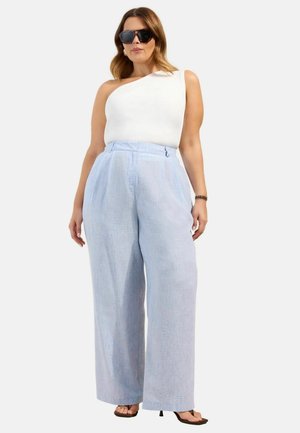 Woman wearing white one-shoulder top, light blue wide-leg pants, sunglasses, and black heeled sandals standing against a plain background.