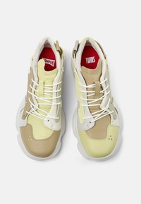 Tan and yellow athletic sneakers with white soles, textured materials, laces, and red branding inside the tongue. Clean, modern silhouette.