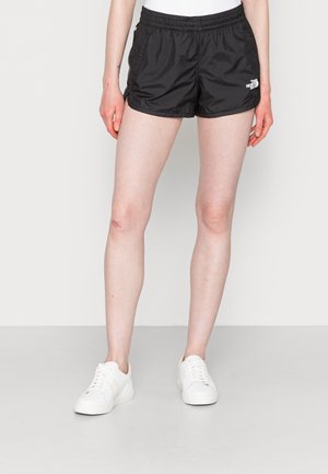 Osoba ubrana w czarne szorty North Face i białe trampki stoi na jednolitym jasnym tle.