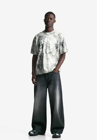 Junger Mann steht in einem marmorierten weißen und grauen übergroßen T-Shirt, weiten abgetragenen schwarzen Jeans und schwarzen Sneakers vor einem weißen Hintergrund.