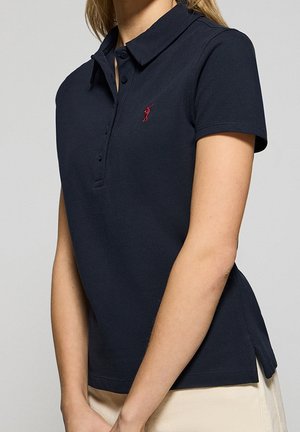 Femme portant un polo à manches courtes bleu marine avec un petit logo brodé rouge sur la poitrine, assorti à un pantalon de couleur claire.