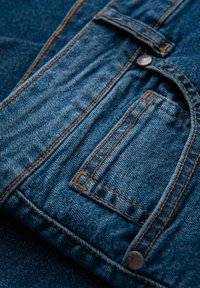 Tissu en denim bleu foncé, présentant un grain subtil, des coutures orange et une petite poche avant avec un bouton métallique.