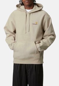 Sudadera con capucha beige hecha de material suave, cuenta con un bolsillo canguro, puños acanalados y un logo bordado en el pecho.