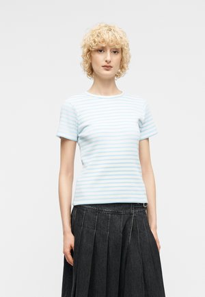 Ξανθό άτομο με σγουρά μαλλιά που φοράει ριγέ κοντομάνικο πουκάμισο σε ανοιχτό γαλάζιο και άσπρο και μαύρη πλισέ φούστα, στέκεται μπροστά σε λευκό φόντο.