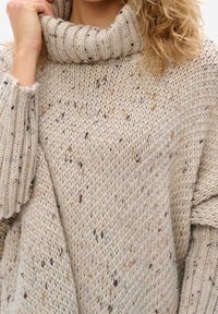 Cabled, turtleneck sweater in a light beige color with scattered dark and light specks. Features wide, ribbed cuffs and a loose fit.