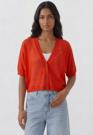 Woman with long dark hair wearing a bright orange short-sleeve buttoned cardigan over a white top and light blue jeans against a plain background.