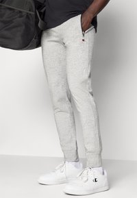 Lichtgrijze joggingbroek met een taps toelopend ontwerp, voorzien van een ritszak en een klein logo aan de zijkant, gecombineerd met witte sneakers.