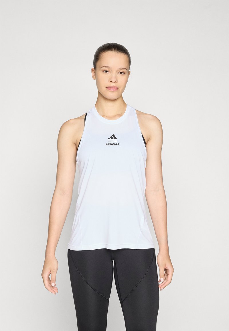 Valge sportlik tankülik, ümmarguse kaelusega, millel on mustad logo aksessuaarid. Kergest materjalist sile tekstuur. Kombineeritud mustade retuusidega.