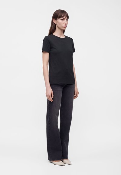 Femme debout portant un t-shirt noir uni à manches courtes, un jean large et foncé, et des chaussures pointues de couleur claire sur fond blanc.