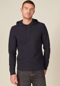 Felpa con cappuccio blu navy, maniche lunghe, tessuto a coste e coulisse. Abbinata a jeans grigio scuro, il modello è in piedi su uno sfondo neutro.