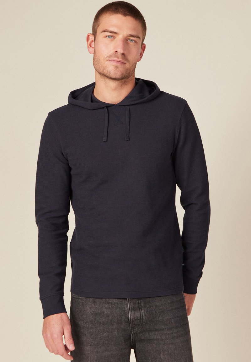 Felpa con cappuccio blu navy, maniche lunghe, tessuto a coste e coulisse. Abbinata a jeans grigio scuro, il modello è in piedi su uno sfondo neutro.