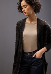 Svart, fluffig cardigan över en beige topp, kombinerad med mörka jeans. Cardiganen har en öppen front och sidofickor.