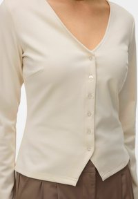 Haut crème à manches longues avec col en V et boutons, associé à un pantalon plissé marron, visible de la poitrine aux hanches.