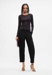 Top negro de manga larga transparente con lunares blancos, combinado con pantalones de pierna ancha negros. La modelo sostiene un clutch burdeos y lleva puestos tacones con tiras.