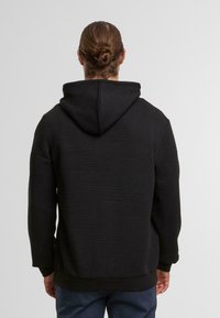 Felpa nera con motivo a zigzag testurizzato, dotata di cappuccio e polsini a costine. Il materiale sembra morbido e caldo, progettato per il comfort.