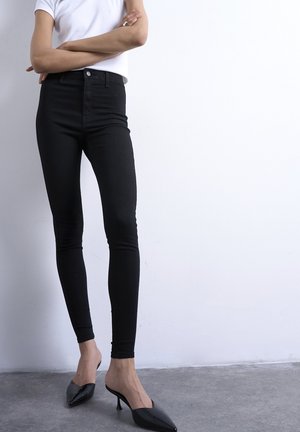 Personne debout, bras croisés, portant une chemise blanche à manches courtes, un pantalon noir moulant et des mules à talons pointues noires sur un sol gris.