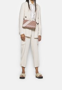 Brązowa torebka kopertówka w deseń, z różowym skośnym paskiem, niesiona przez osobę w jasnobeżowym garniturze i sandałach. Płaski, prostokątny kształt.