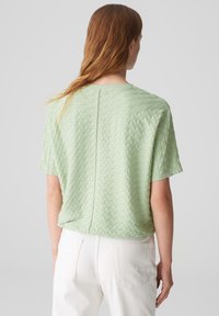 Haut à manches courtes de couleur vert pâle, texturé, avec une coupe ample et une couture arrière droite, porté avec un jean blanc. Motif texturé sur l'ensemble.