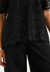 Schwarzes Spitzenoberteil mit floralem Muster und gewelltem Saum, kombiniert mit schwarzen Jeans. Details sind fransige Kanten und transparente Textur.