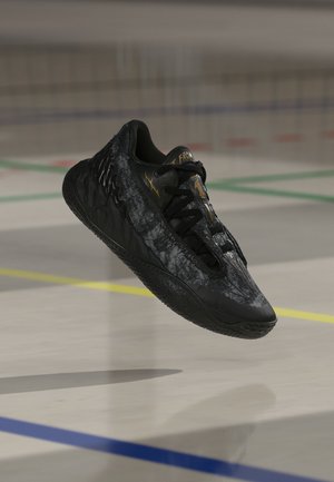 Schwarzer Sportschuh mit strukturierten Details und Schnürsenkeln, der über einem Hallensportplatz mit farbigen Linien schwebt.
