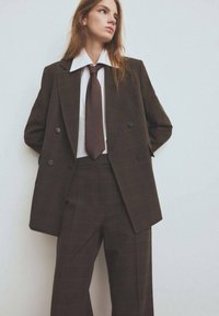 Kobieta w brązowym w kratę garniturze z szerokimi spodniami, białej koszuli i brązowym krawacie, stojąca z rękami za plecami na jednolitym tle.