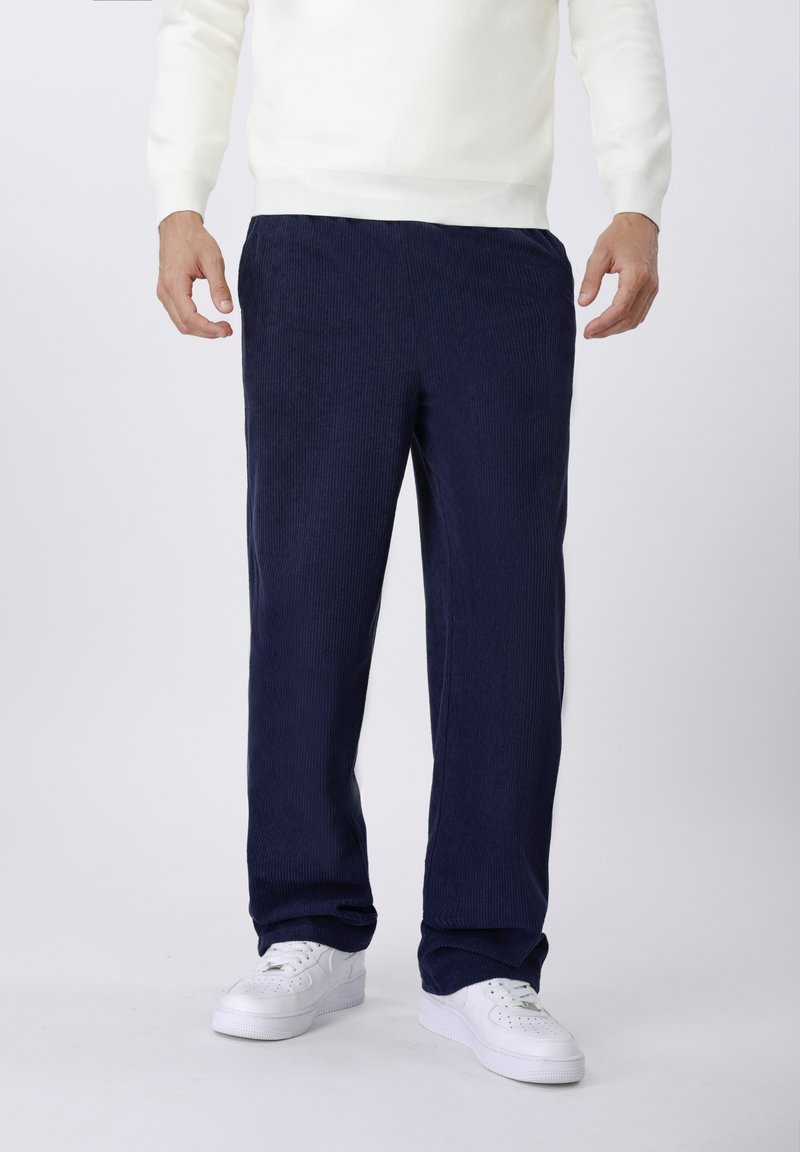 Pantalon en velours côtelé bleu marine à coupe droite avec ceinture, présentant des rayures verticales texturées. Associé à des baskets blanches et un pull léger.
