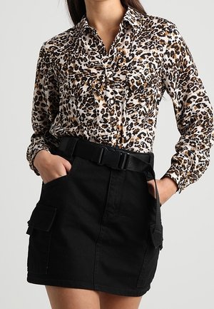 Vrouw draagt een longsleeve met luipaardprint, gestopt in een zwarte rok met hoge taille, met een dikke zwarte riem en cargozakken.