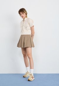 Beige kortermet skjorte med to lommer, sammen med en khaki plisserte skjørt. Bruker kremfargede sneakers med brune og oransje detaljer.