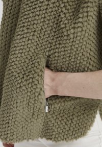 Olivgrüne, flauschige Stoffjacke mit einer strukturierten, knubbeligen Oberfläche; verfügt über eine mit Reißverschluss ausgestattete Tasche an der Seite für praktischen Stauraum.