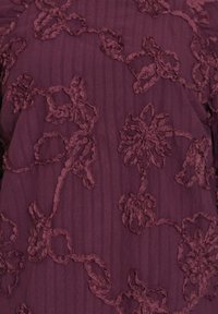 Burgundy blus med texturerad blommönsterbroderi med upphöjda detaljer, placerad på en slät randig tygbakgrund.